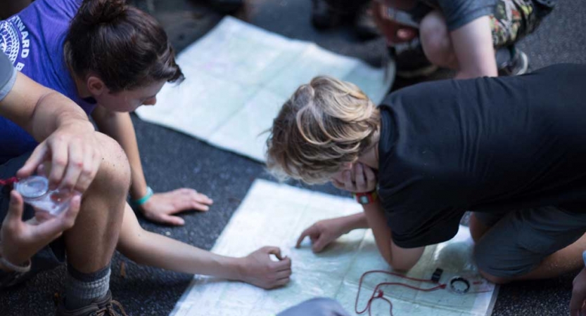 Two students point at a map that is laid out on the ground. 